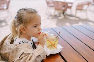 Little girl drinks juice from a glass through a straw while sitting at the table. Side view photo