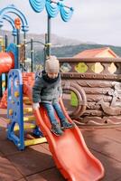 Little smiling girl in mittens slides down a slide on the playground photo