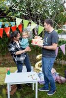 Little smiling girl sits in the arms of her mother standing next to her father with a birthday cake in his hands photo