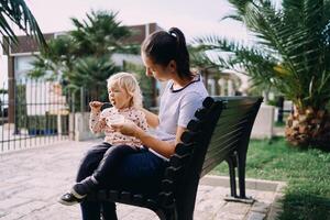 Little girl with a spoon eats porridge from a lunchbox in the hands of her mom sitting on a bench photo