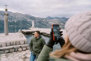Woman takes a picture with a smartphone of a man standing on a pier near a column with a cross. Back view photo