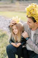 Smiling dad in a wreath of yellow leaves puts autumn leaves on the head of a little girl sitting on his lap photo