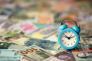 Small alarm clock on many banknotes of different currency. Background of time and money photo