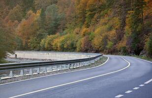 Road in autumn forest at sunset in Carpathian mountains, Ukraine. Beautiful mountain roadway with orange tress and high rocks. Landscape with empty highway through the woods in fall photo