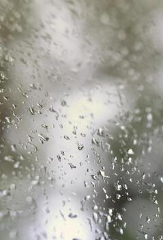 A photo of rain drops on the window glass with a blurred view of the blossoming green trees. Abstract image showing cloudy and rainy weather conditions