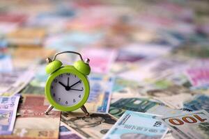 Small alarm clock on many banknotes of different currency. Background of time and money photo