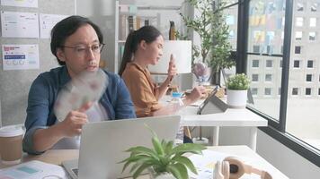 Two Asian workers are unhappy working with hot temperatures at casual office in summer season, uncomfortable with no air conditioner, using fans and drinking water, sitting at desks, and sweating. video