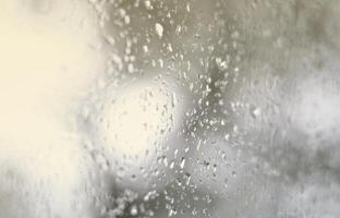A photo of rain drops on the window glass with a blurred view of the blossoming green trees. Abstract image showing cloudy and rainy weather conditions
