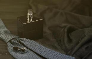 A bottle of mens cologne and cufflinks with blue tie lie on a black luxury fabric background on a wooden table. Mens accessories photo