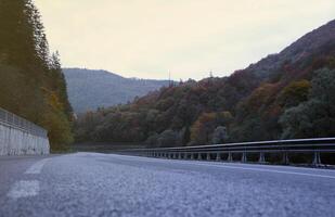 Road in autumn forest at sunset in Carpathian mountains, Ukraine. Beautiful mountain roadway with orange tress and high rocks. Landscape with empty highway through the woods in fall photo