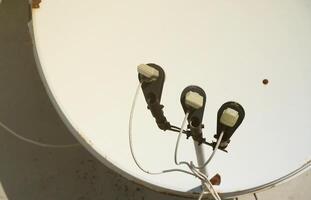 White satellite dish with three converters mounted on residental building rooftop concrete wall. Satellite television photo