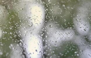 A photo of rain drops on the window glass with a blurred view of the blossoming green trees. Abstract image showing cloudy and rainy weather conditions