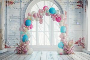 A large archway of balloons is set up in a room with a window. The archway is made up of pink, blue, and white balloons photo