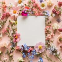 Square blank card surrounded by pastel colored flowers on a light pink background in a top view photo