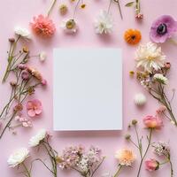 Square blank card surrounded by pastel colored flowers on a light pink background in a top view photo