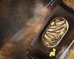 A open tin of Baltic sprats with spices, a fork, a bottle of dark beer and rye bread on brown background photo