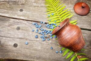 A clay pot with scattered blueberries and fern leave on a wooden table photo
