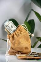 A brown cloth bag with a drawstring sits on a white surface. The bag is filled with US dollar bills and a few coins are scattered on the table photo