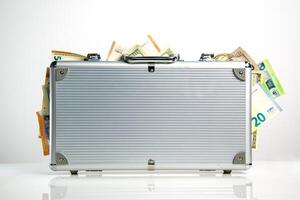 A silver aluminum briefcase with a slightly open latch and a few protruding bills of various currencies photo