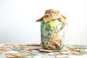A glass jar filled with various denominations of US dollar and euro bills. The jar is covered with a brown paper bag and surrounded by scattered bills photo