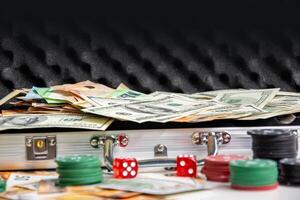 A metal case with a black interior filled with US dollar bills and euro bills, and stacks of red and green poker chips are scattered on a table in front of the case, with two red dice on the table photo