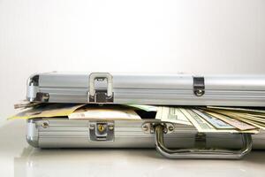 A close-up of an open silver aluminum case with cash spilling out from inside. The case is on a white surface with the cash slightly overlapping the edge photo
