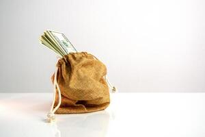 A brown canvas bag with a drawstring sits on a white surface. Several US dollar bills are sticking out of the top of the bag photo