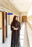 A Muslim student wearing traditional clothes standing and hold Quran on walkway in her school building with sun flare background. photo