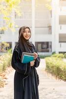 Cheerful Asian Muslim wearing traditional clothes stand and holding textbook on walkway on her school building with sun flare background. photo