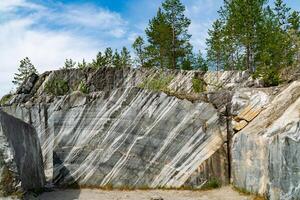 Scenic view of a marble quarry with distinct striations and pine trees in the background. The image highlights the contrast between the natural stone formations and the greenery under a bright blue sky. photo