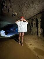 A woman wearing an oversized white t-shirt and a red helmet explores a dimly lit cave, looking up at the stalactites and cave formations with a curious expression. photo