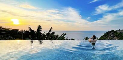 A woman enjoys a serene moment in an infinity pool overlooking the ocean at sunset. The lush tropical scenery and peaceful ambiance create a perfect setting for themes of relaxation, luxury, and travel. photo
