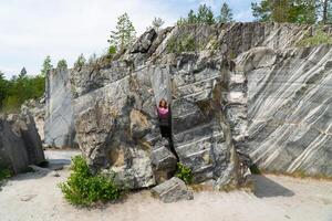 A woman standing in a marble quarry surrounded by unique rock formations. The image captures the natural beauty of the quarry with its distinctive marble striations and the greenery in the background photo
