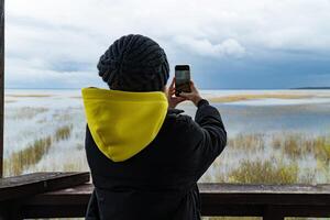 A woman in a yellow hoodie and black jacket stands on a tower, capturing a flock of birds nesting on the horizon with her smartphone. The scene highlights the beauty of wildlife and the excitement of bird watching photo