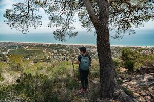 Hiker with a backpack and mountain sticks, contemplates the Mediterranean coast from above and below a tree. photo