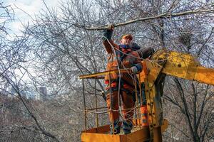 Dnepr, Ukraine - 03.20.2024 Municipal service workers stand with a chainsaw in a crane basket and trim dangerous trees photo