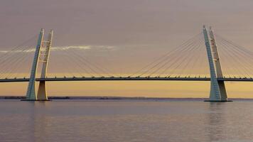 A breathtaking view of a modern cable-stayed bridge silhouetted against a warm sunset sky. The serene waters below reflect the bridge's elegant structure, creating a perfect blend of nature and architecture. video