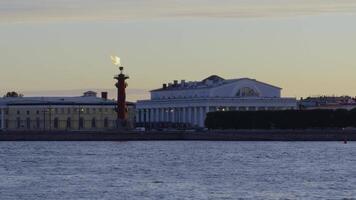 Scenic view of the iconic Rostral Column with a flame at its top, set against the backdrop of historical buildings in Saint Petersburg during sunset. The Neva River in the foreground enhances the tranquil atmosphere. video
