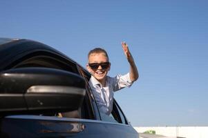 A cute boy in sunglasses looked out of the car window waving his hand with a laugh photo