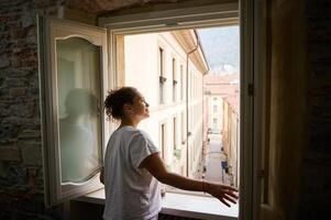 Side view. Smiling happy young woman in white pajamas, opening windows in the living room, contemplating the beautiful touristic city of Como in Lombardy, with red roof tiles of cozy houses. Italy photo