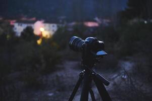 A digital camera with black mockup touch screen placed on tripod by a travel photographer hiker, capturing the beautiful view of a city in mountains at dawn. The concept of World photography Day photo