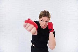 Athletic woman fighter with red tapes around her fists, punching forward while practicing boxing exercises over white isolated background. Copy space for text. People, sport and martial art concept photo