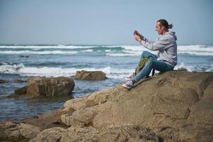 Man sitting on rocky shoreline taking photo with smartphone at the beach