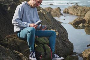Man relaxing on rocky coast while enjoying a hot drink from a thermos bottle photo