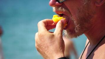 Man eating peach. Bearded man in sunglases eating peach on the beach. Happy man is smiling and resting at sea. Bearded guy enjoying fruits on summer vacation. Vegetarian and healthy lifestyle concept. video