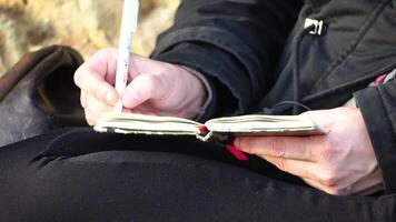 Woman writes in handbook. Close up. Young inspired woman is sitting on seashore and writing with pen in notebook. Lady writing notes in copybook. Freelancer, blogger working on notebook on the beach. video