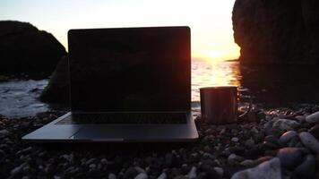 Silhouette of laptop and cup on the beach over sea sunset. Sparkling sea water on background. Freelancer, blogger working on notebook on the beach. video