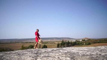 woman in a red dress and straw hat standing on a rock, enjoying the fresh air. Embracing freedom and beauty of nature. Happy lady on top of mountain with raised hands while wind is blowing her dress. video