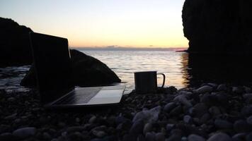 Silhouette of laptop and cup on the beach over sea sunset. Sparkling sea water on background. Freelancer, blogger working on notebook on the beach. video