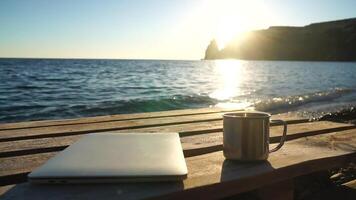 Silhouette of laptop and cup on the beach over sea sunset. Sparkling sea water on background. Freelancer, blogger working on notebook on the beach. video